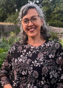 A head-and-shoulders portrait of a smiling woman with gray hair and glasses. She is wearing a black, floral-patterned blouse and large hoop earrings. The background is a slightly blurred outdoor scene with greenery and a wooden fence.